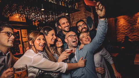 A stock image of a group of friends having fun at a bar with a man taking a selfie from his cell phone -- this man recently got Blepharoplasty surgery and used LidLift Goggle to recover